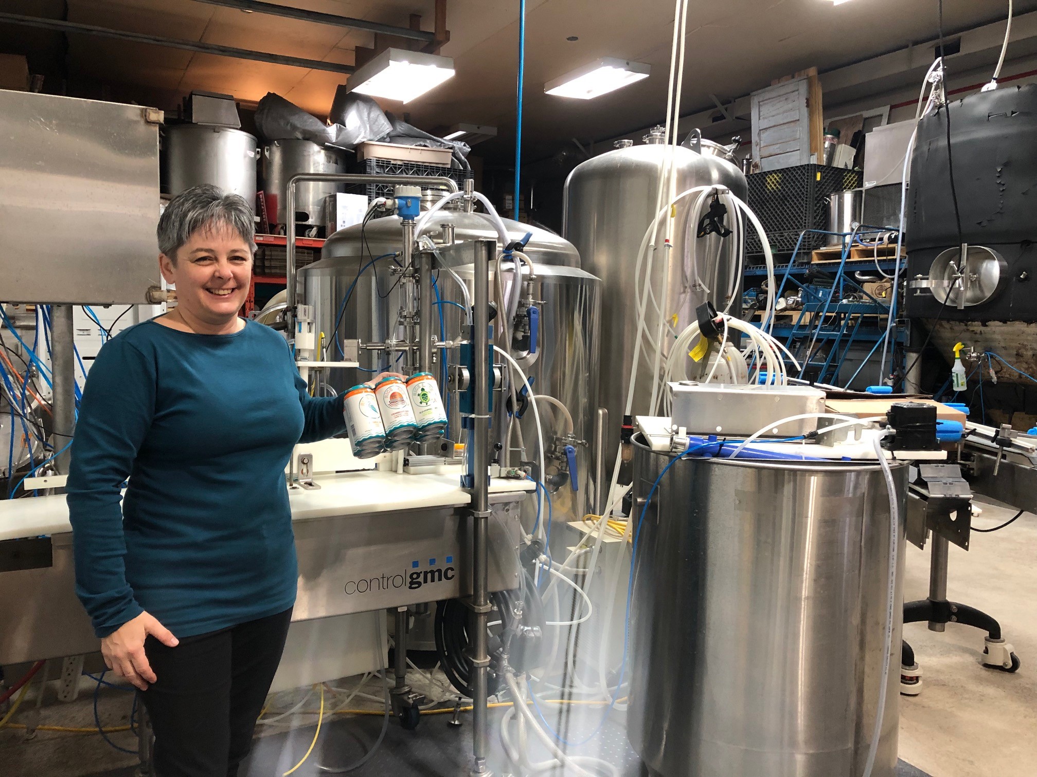 Business owner of Rice Lake Hard Cider smiles, holding case of cider in front of equipment
